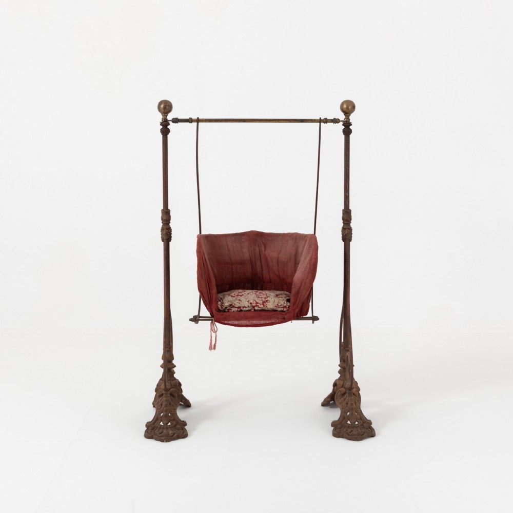 Victorian cast iron freestanding child swing featuring scrolled feet, brass finials, and a red fabric-upholstered suspended seat. View III