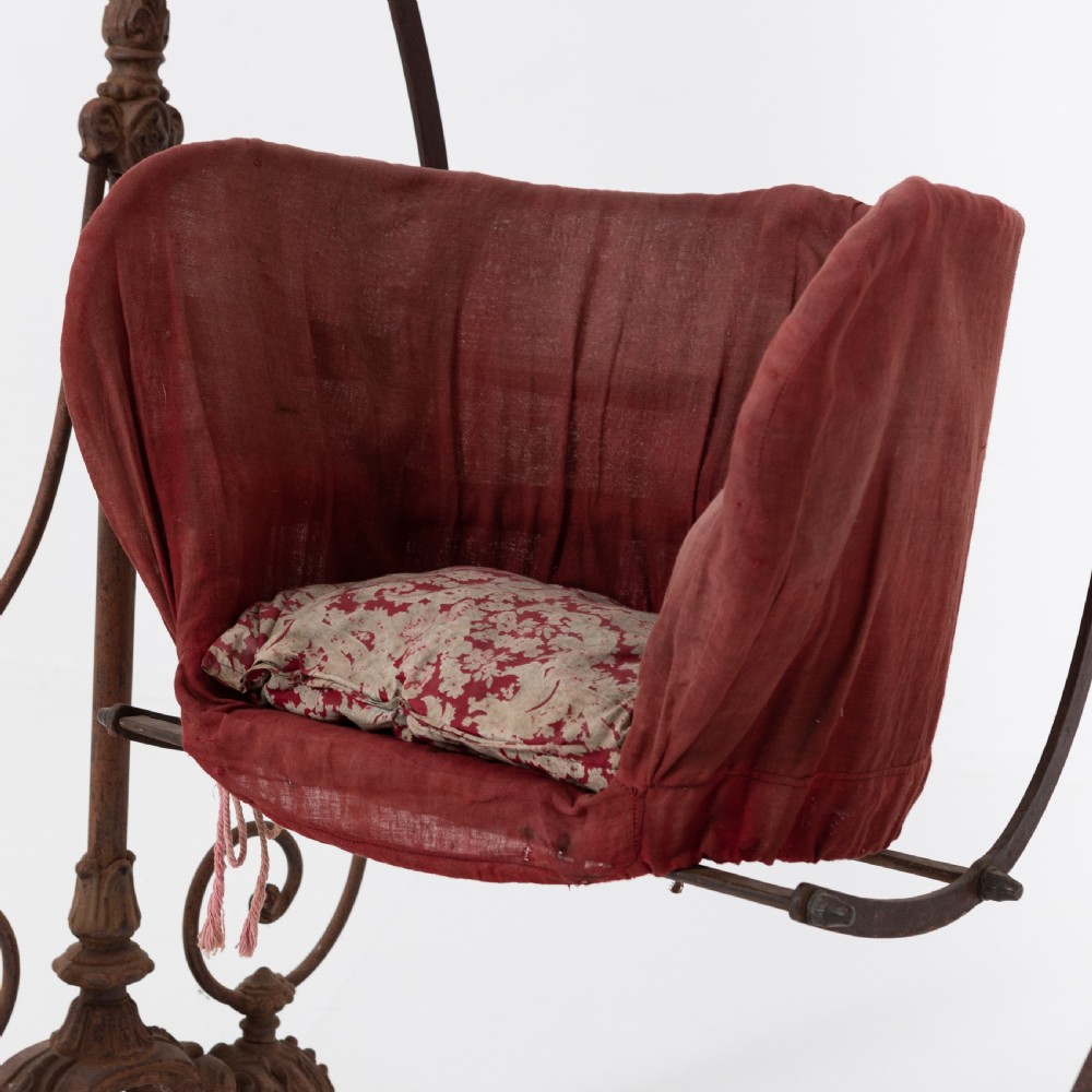 Victorian cast iron freestanding child swing featuring scrolled feet, brass finials, and a red fabric-upholstered suspended seat. View V