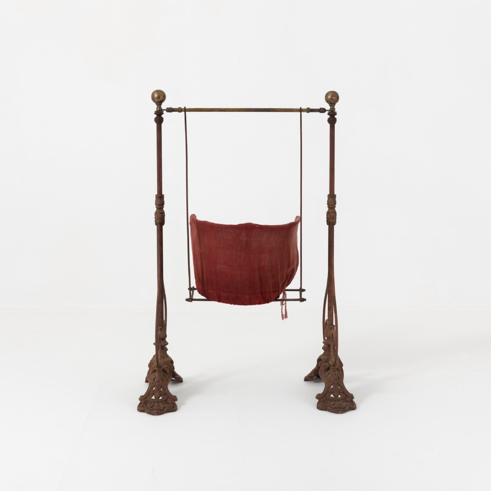 Victorian cast iron freestanding child swing featuring scrolled feet, brass finials, and a red fabric-upholstered suspended seat. View VI