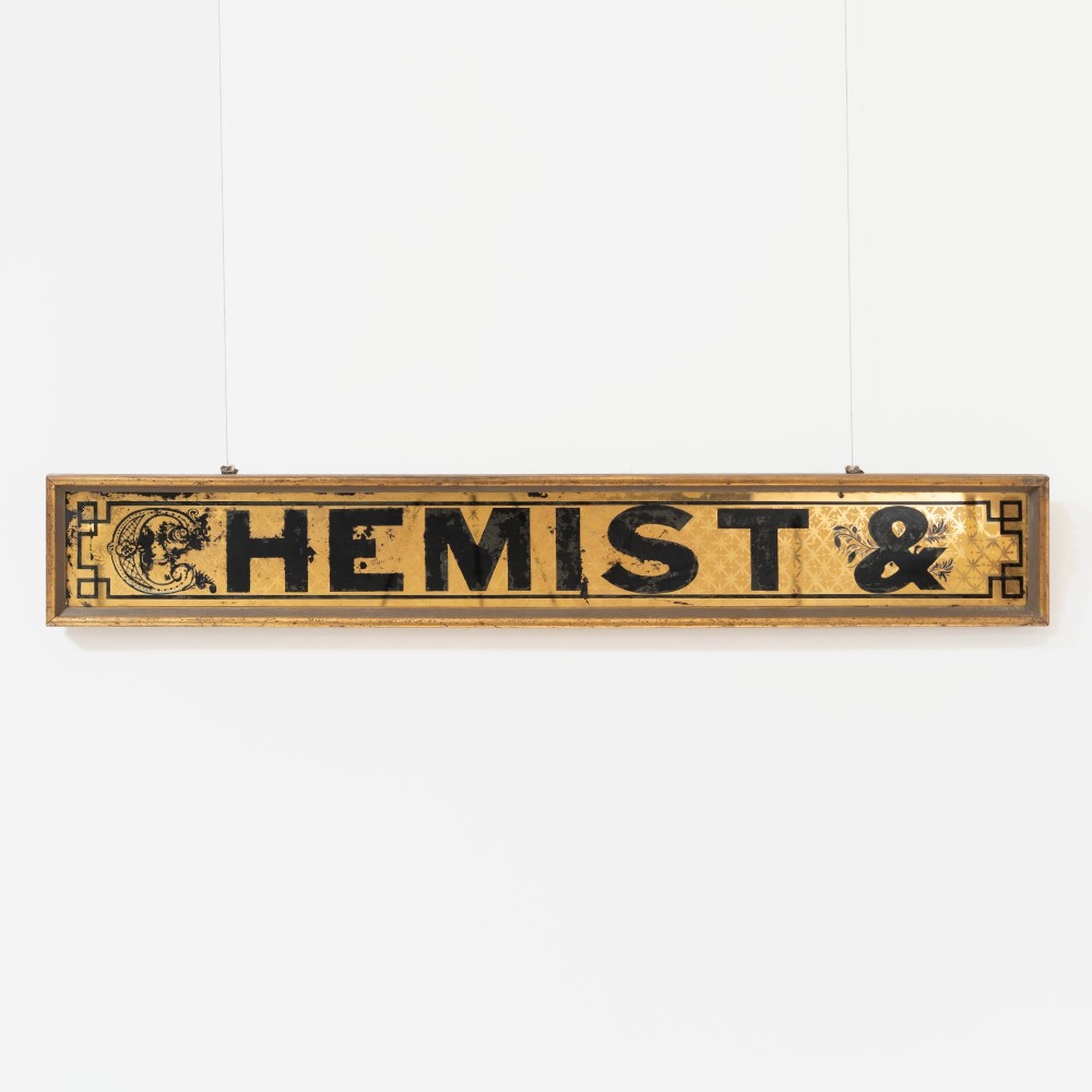 A long rectangular late Victorian verre eglomise glass shop sign framed in gold timber, displaying bold black lettering reading CHEMIST & on a gilded background.