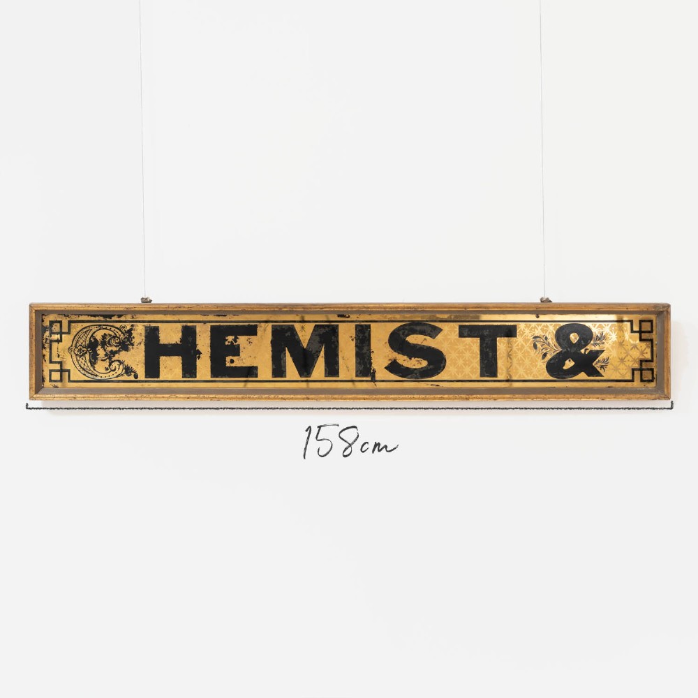 A long rectangular late Victorian verre eglomise glass shop sign framed in gold timber, displaying bold black lettering reading CHEMIST & on a gilded background. View II