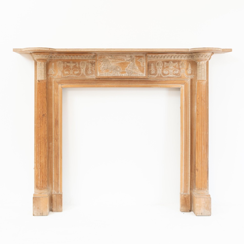 A Victorian stripped pine fireplace surround featuring fluted side columns, a stepped mantel shelf, and a central carved panel depicting a chariot scene.