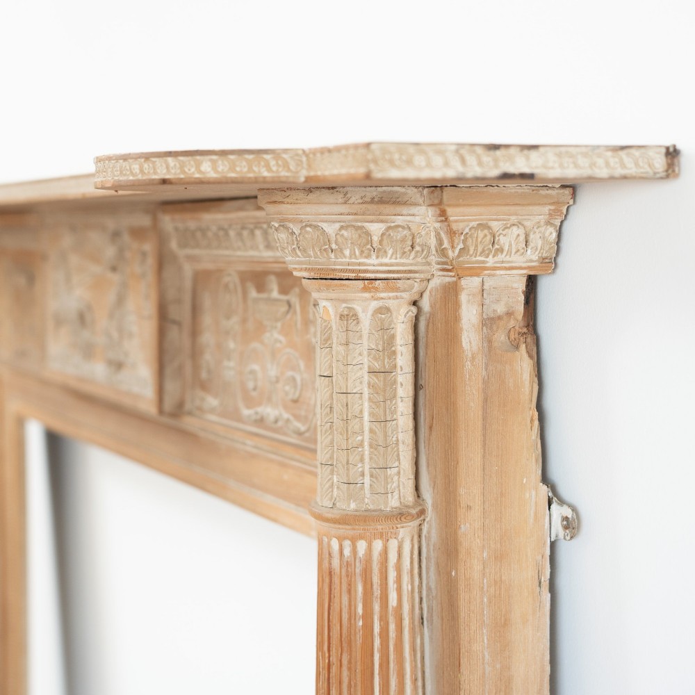 A Victorian stripped pine fireplace surround featuring fluted side columns, a stepped mantel shelf, and a central carved panel depicting a chariot scene. View V