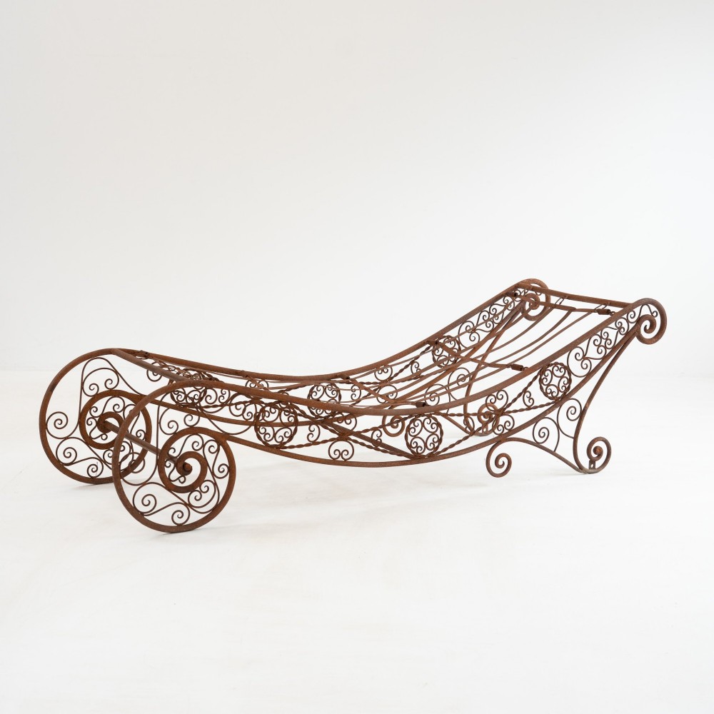 A nineteenth-century French wrought iron garden daybed featuring scrollwork, a contoured reclining frame, and decorative front wheels with a weathered brown patina.
