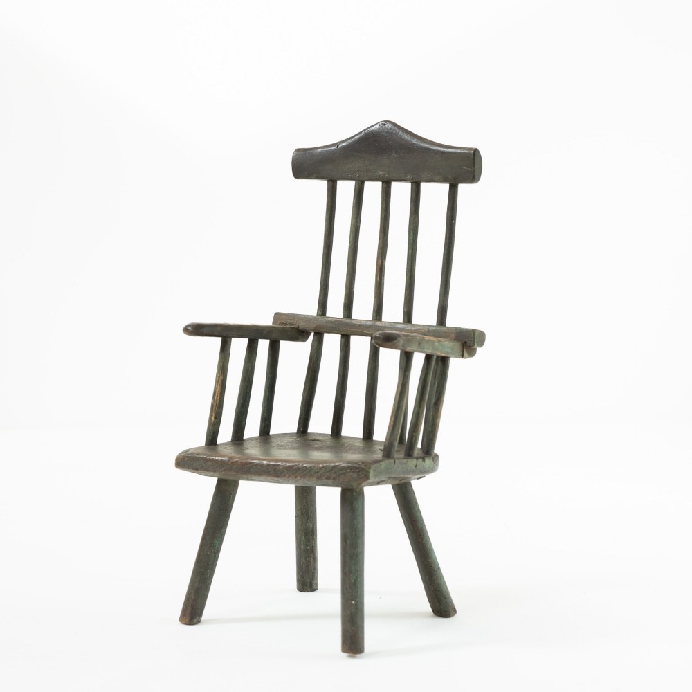 A 19th-century primitive childs stick-back armchair crafted from dark patinated timber with splayed legs and a crest rail.