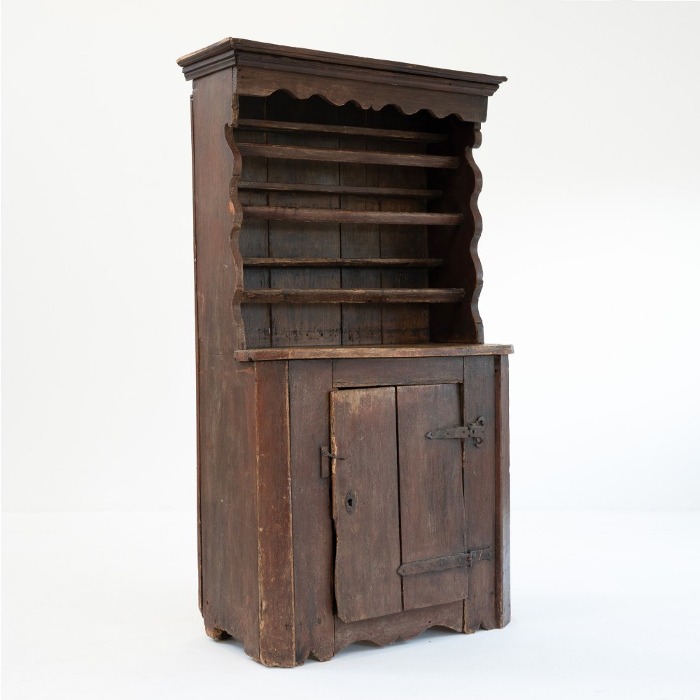 A late eighteenth-century dark timber Irish kitchen dresser featuring open plate shelves with scalloped sides above a single-door cupboard base with wrought iron hinges.
