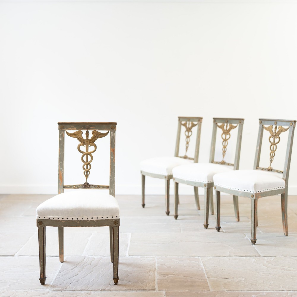 A set of four 19th-century Northern European Neoclassical side chairs featuring distressed grey paintwork, off-white linen upholstery, and gilded caduceus motifs in the open back frames.