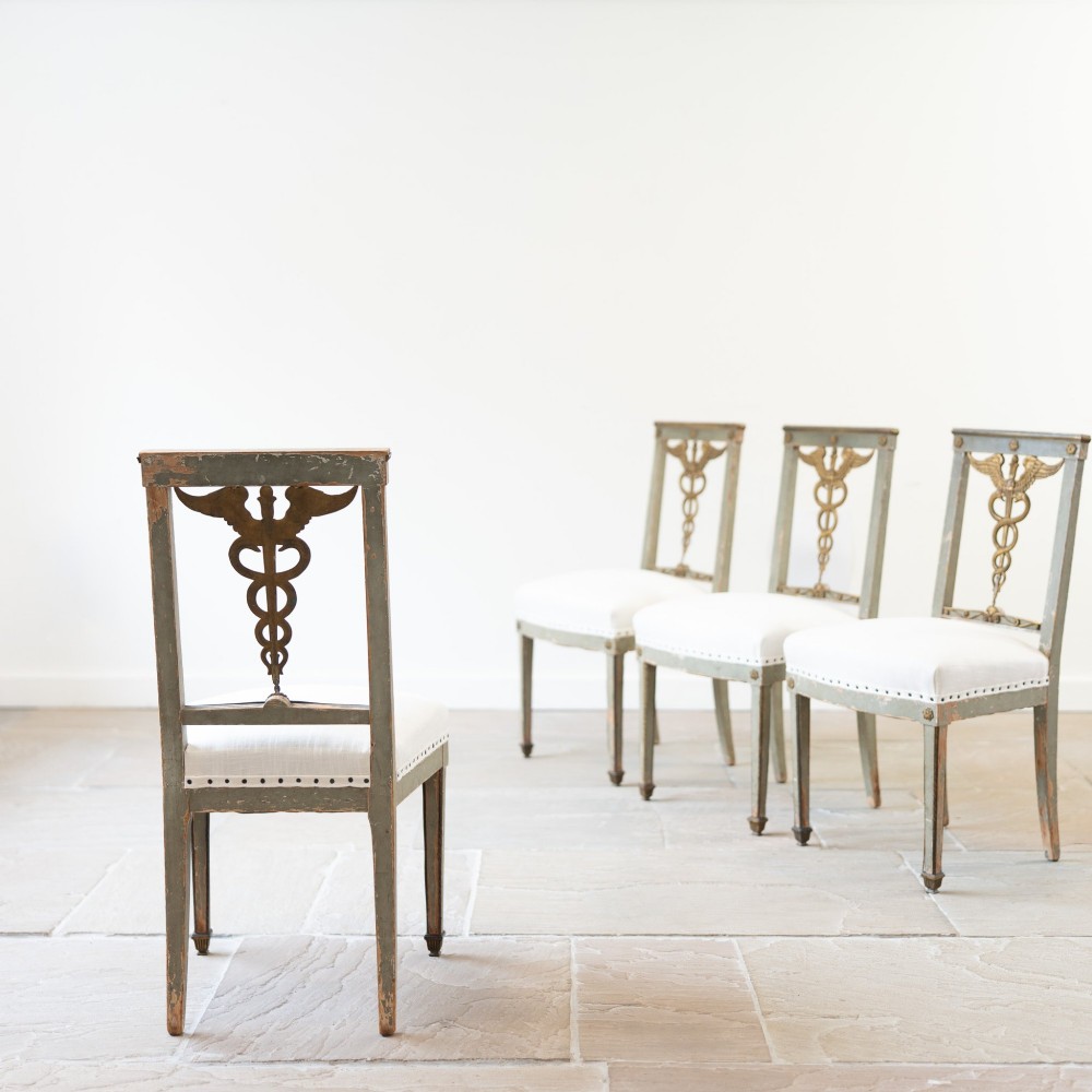 A set of four 19th-century Northern European Neoclassical side chairs featuring distressed grey paintwork, off-white linen upholstery, and gilded caduceus motifs in the open back frames. View III