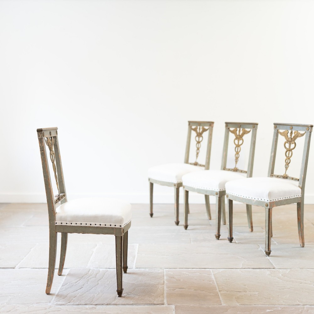 A set of four 19th-century Northern European Neoclassical side chairs featuring distressed grey paintwork, off-white linen upholstery, and gilded caduceus motifs in the open back frames. View IV