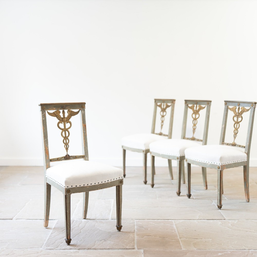A set of four 19th-century Northern European Neoclassical side chairs featuring distressed grey paintwork, off-white linen upholstery, and gilded caduceus motifs in the open back frames. View V