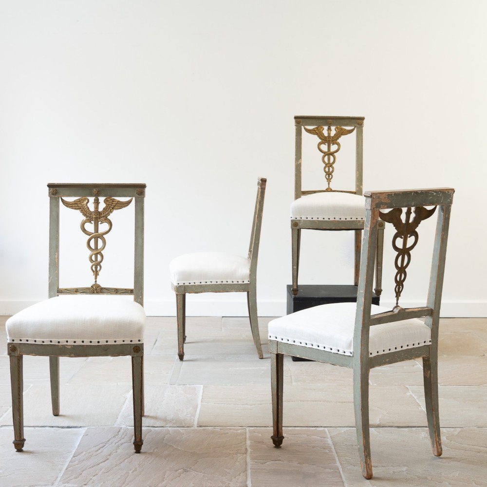 A set of four 19th-century Northern European Neoclassical side chairs featuring distressed grey paintwork, off-white linen upholstery, and gilded caduceus motifs in the open back frames. View VI