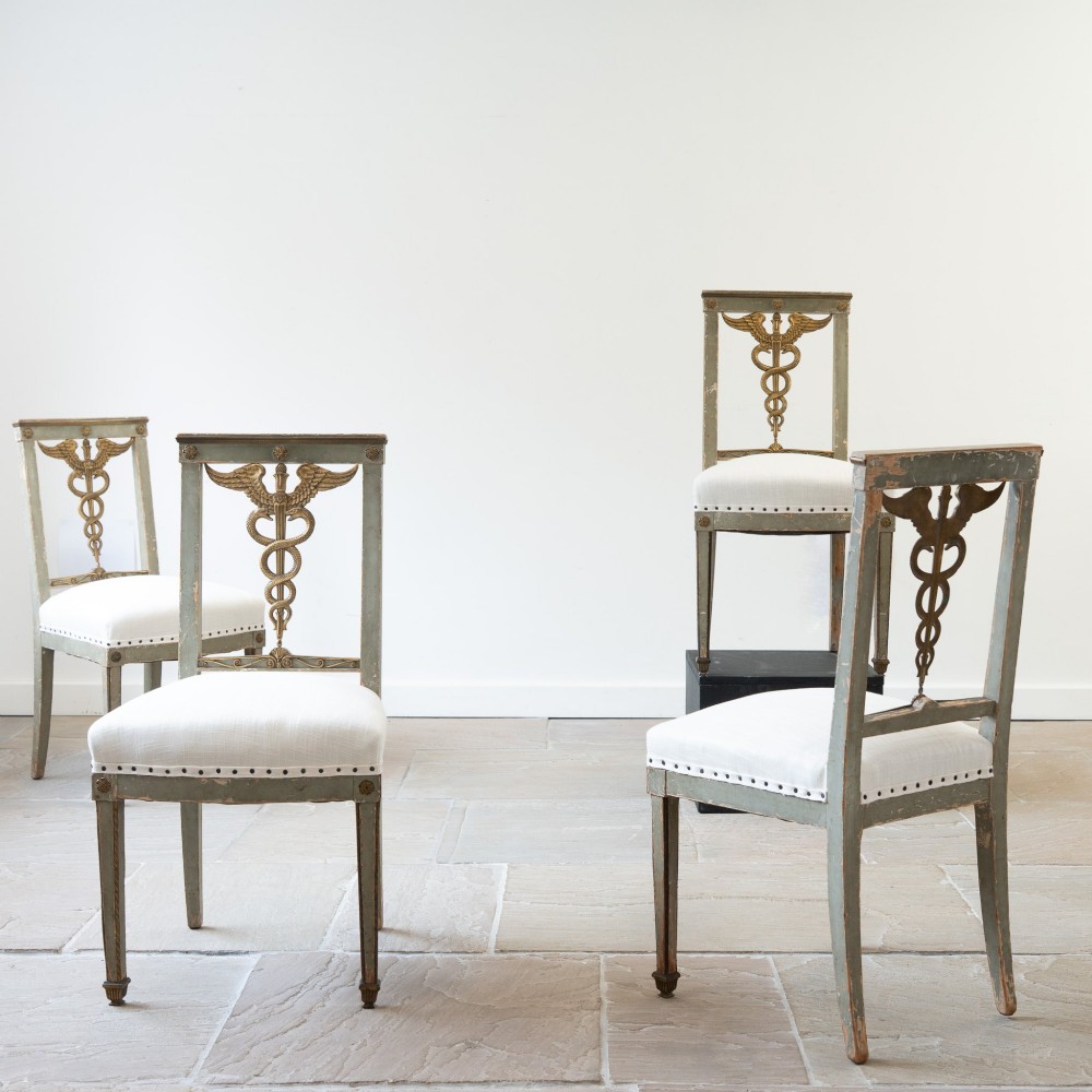 A set of four 19th-century Northern European Neoclassical side chairs featuring distressed grey paintwork, off-white linen upholstery, and gilded caduceus motifs in the open back frames. View VII