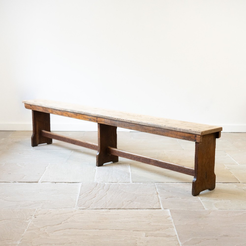 A long English oak refectory bench featuring a rectangular seat supported by three slab legs connected by a low central stretcher.