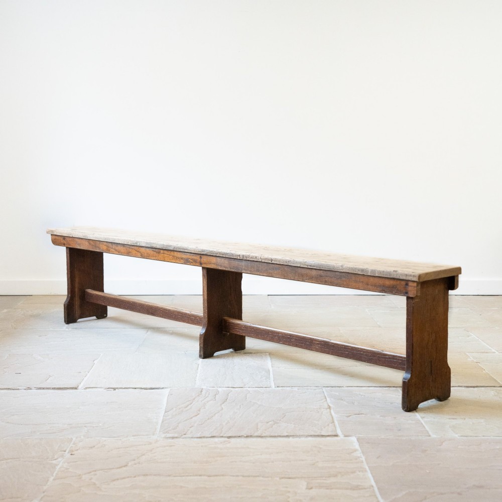 A long English oak refectory bench featuring a rectangular seat supported by three slab legs connected by a low central stretcher. View V