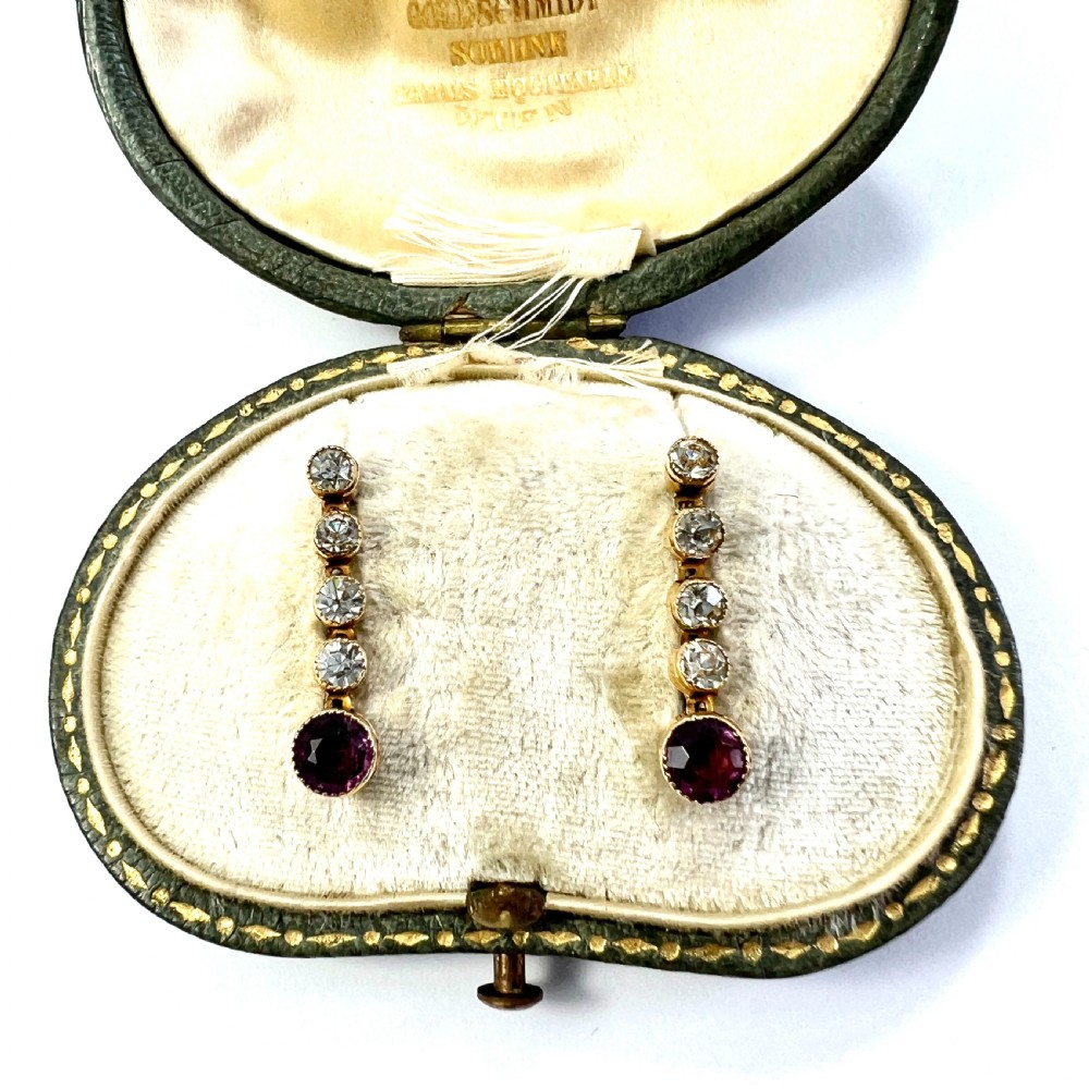 A pair of Edwardian 9ct yellow gold drop earrings with linear clear stones and round purple amethyst terminals set in a green fitted display box.