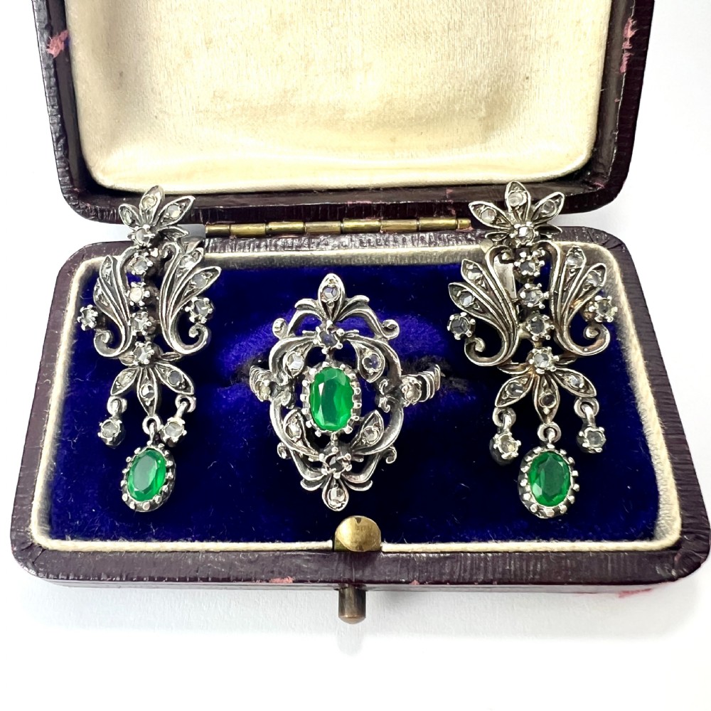 A vintage silver jewellery set featuring a pair of foliate drop earrings and a matching ring, each set with green paste emeralds and clear glass stones.