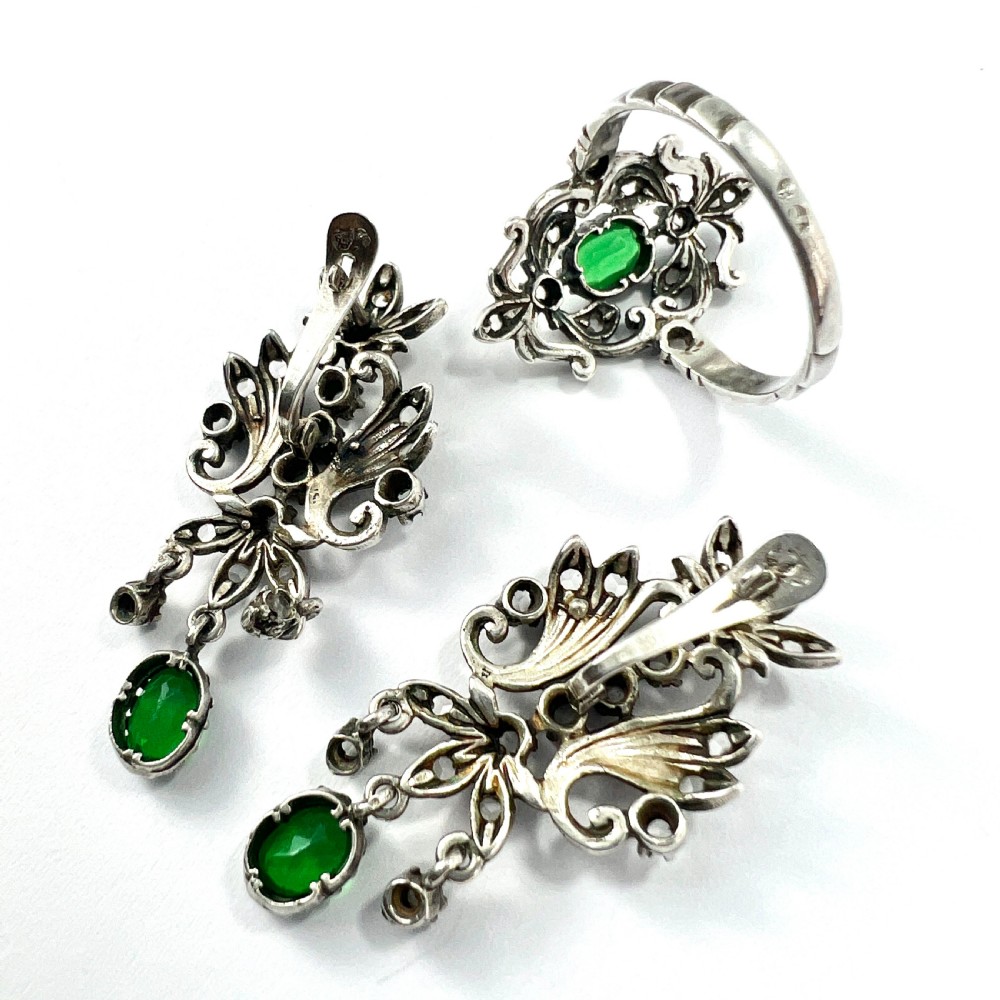 A vintage silver jewellery set featuring a pair of foliate drop earrings and a matching ring, each set with green paste emeralds and clear glass stones. View VI