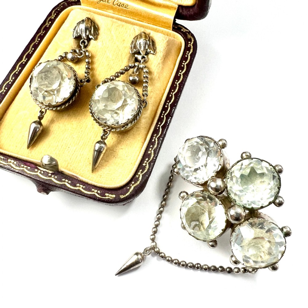 An antique silver demi-parure featuring round clear foiled paste stones in closed collet settings, comprising dangle earrings with cone drops and a matching cluster brooch with a draped beaded chain.