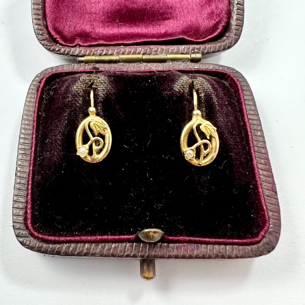 A pair of 9ct yellow gold Art Nouveau dormeuse earrings featuring an oval frame enclosing a botanical scroll motif with a seed pearl accent.
