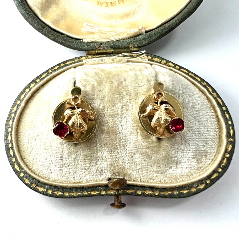 A pair of Victorian 18ct gold stud earrings featuring textured vine leaf motifs and circular red ruby accents set on round gold discs.