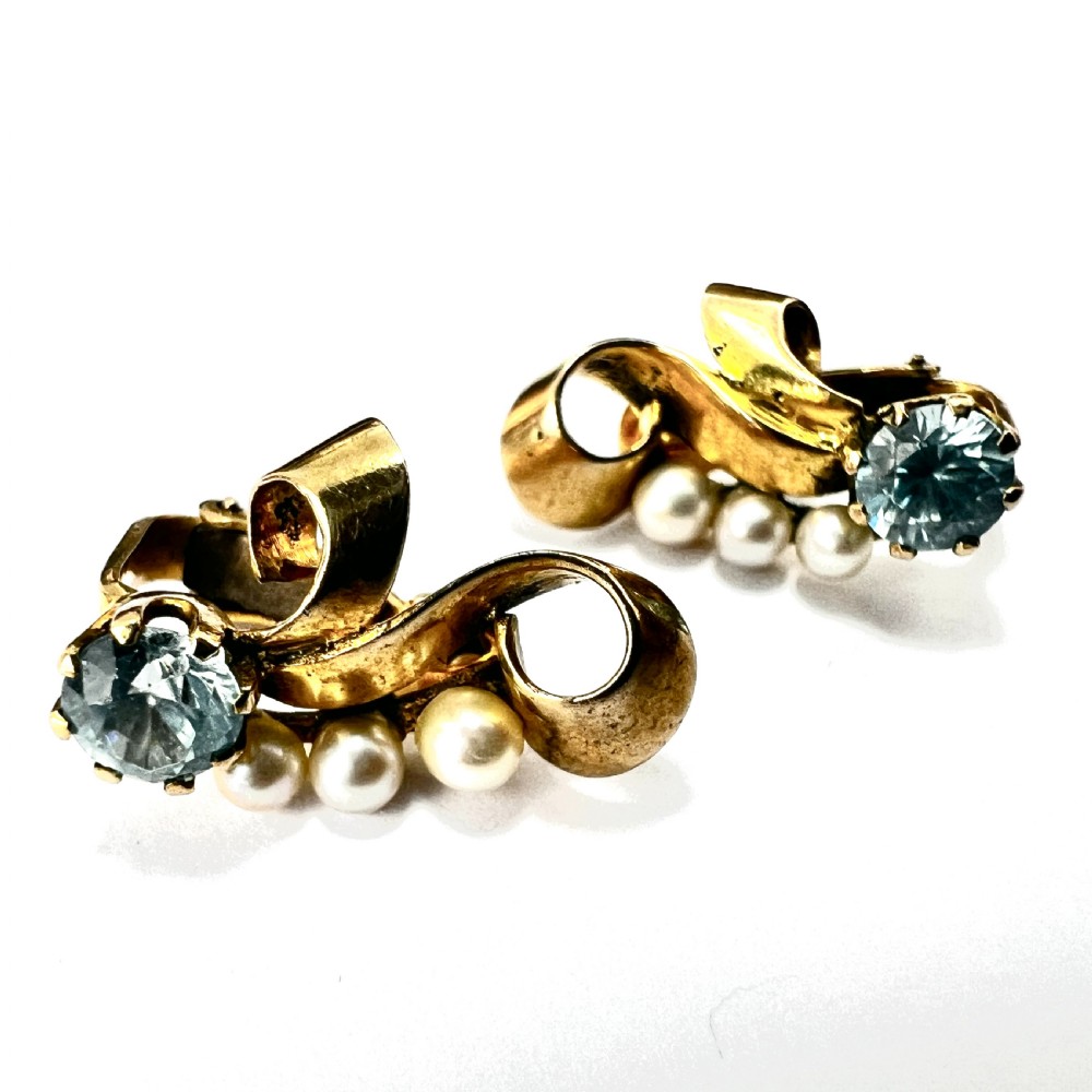 A pair of 1940s 9ct yellow gold ear clips featuring faceted round blue zircons and graduated pearls set against curved ribbon scrollwork. View V