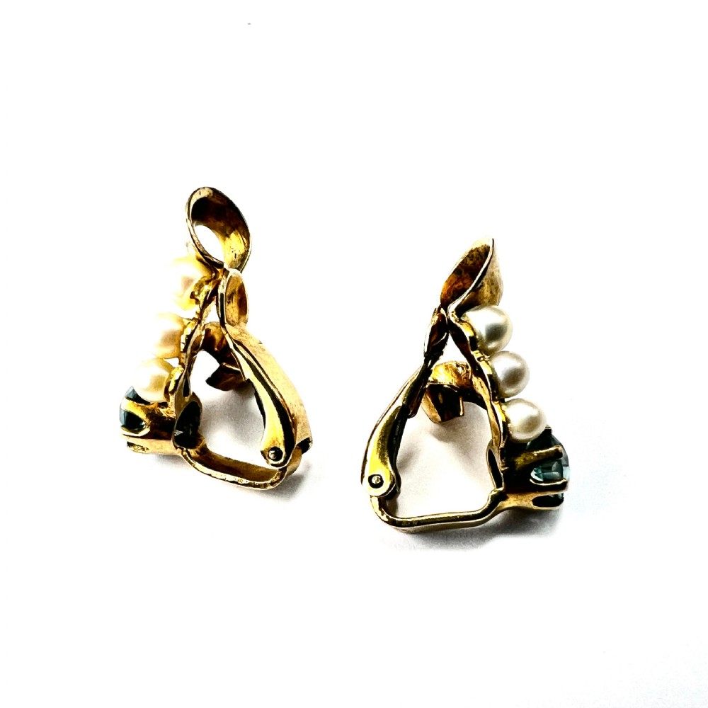 A pair of 1940s 9ct yellow gold ear clips featuring faceted round blue zircons and graduated pearls set against curved ribbon scrollwork. View VI
