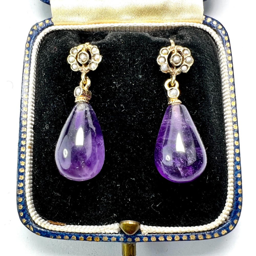 A pair of Victorian gold drop earrings featuring teardrop cabochon amethysts suspended from pearl-set floral clusters inside a fitted velvet box.