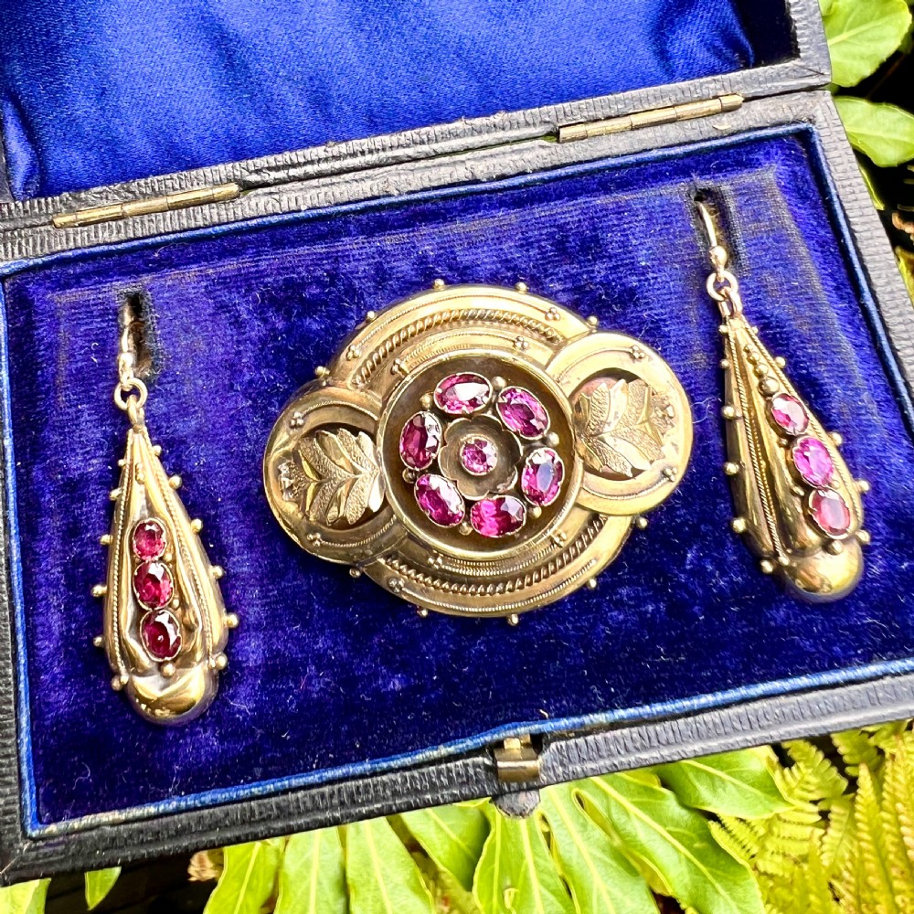 A Victorian gold demi parure set featuring an ornate oval brooch and matching drop earrings inset with amethysts, presented inside a blue velvet-lined case. View X
