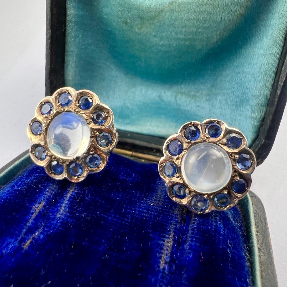 A pair of circular gold screwback earrings, each featuring a round cabochon moonstone surrounded by a halo of small blue sapphires.