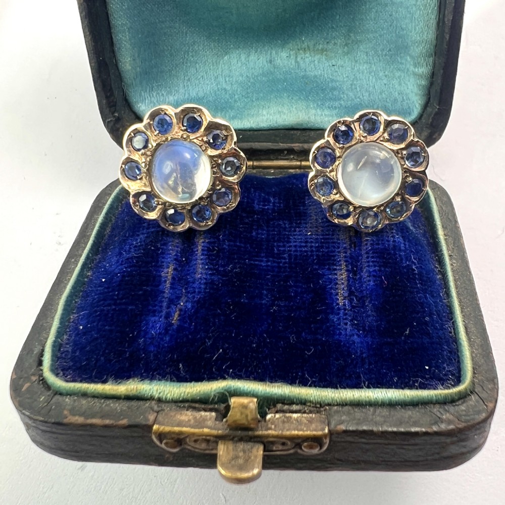 A pair of circular gold screwback earrings, each featuring a round cabochon moonstone surrounded by a halo of small blue sapphires. View II