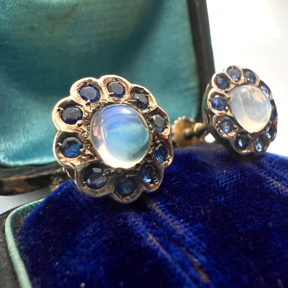A pair of circular gold screwback earrings, each featuring a round cabochon moonstone surrounded by a halo of small blue sapphires. View III
