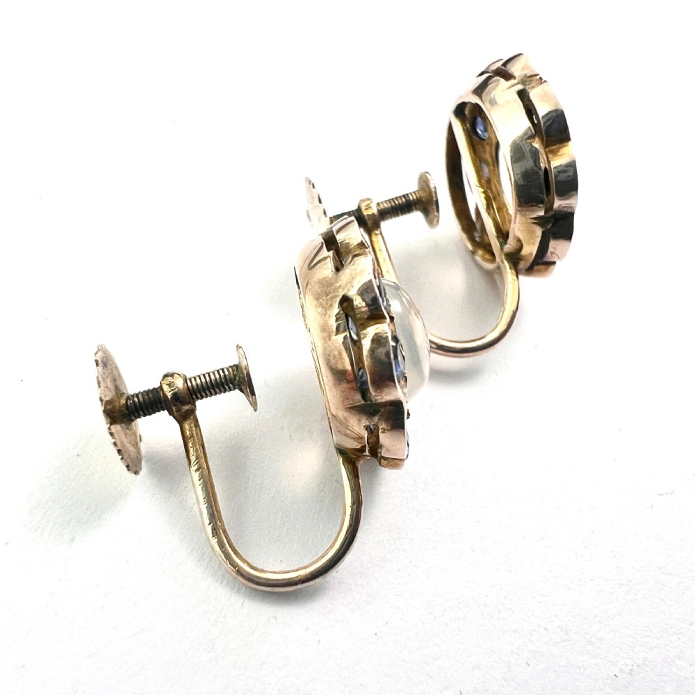 A pair of circular gold screwback earrings, each featuring a round cabochon moonstone surrounded by a halo of small blue sapphires. View V