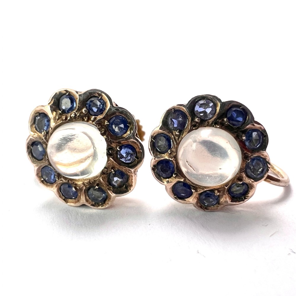 A pair of circular gold screwback earrings, each featuring a round cabochon moonstone surrounded by a halo of small blue sapphires. View VI