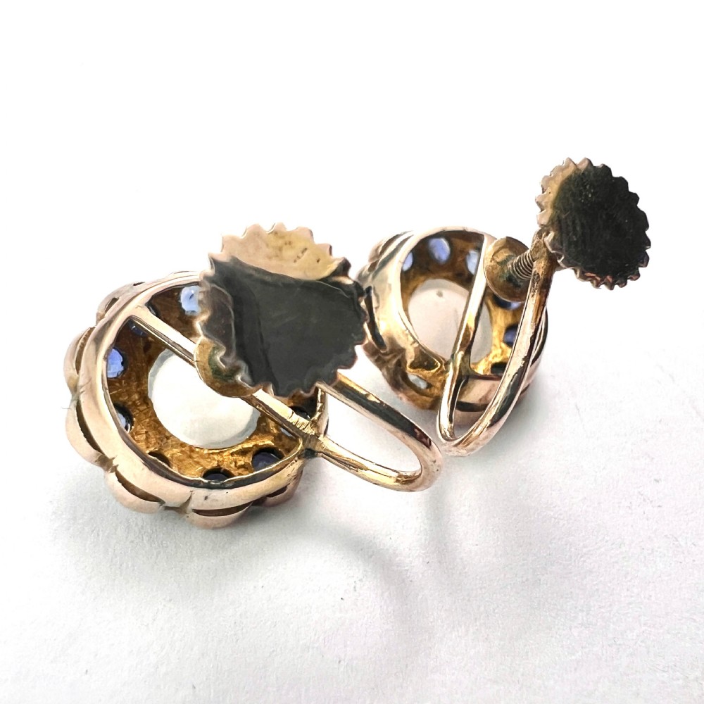 A pair of circular gold screwback earrings, each featuring a round cabochon moonstone surrounded by a halo of small blue sapphires. View VII