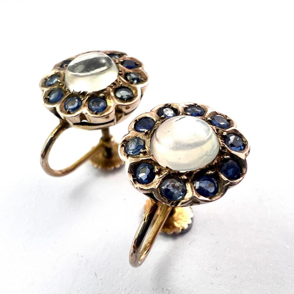 A pair of circular gold screwback earrings, each featuring a round cabochon moonstone surrounded by a halo of small blue sapphires. View VIII