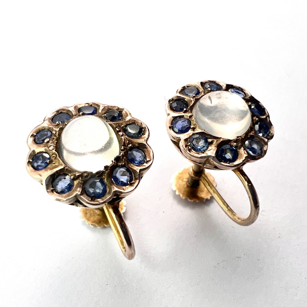 A pair of circular gold screwback earrings, each featuring a round cabochon moonstone surrounded by a halo of small blue sapphires. View IX