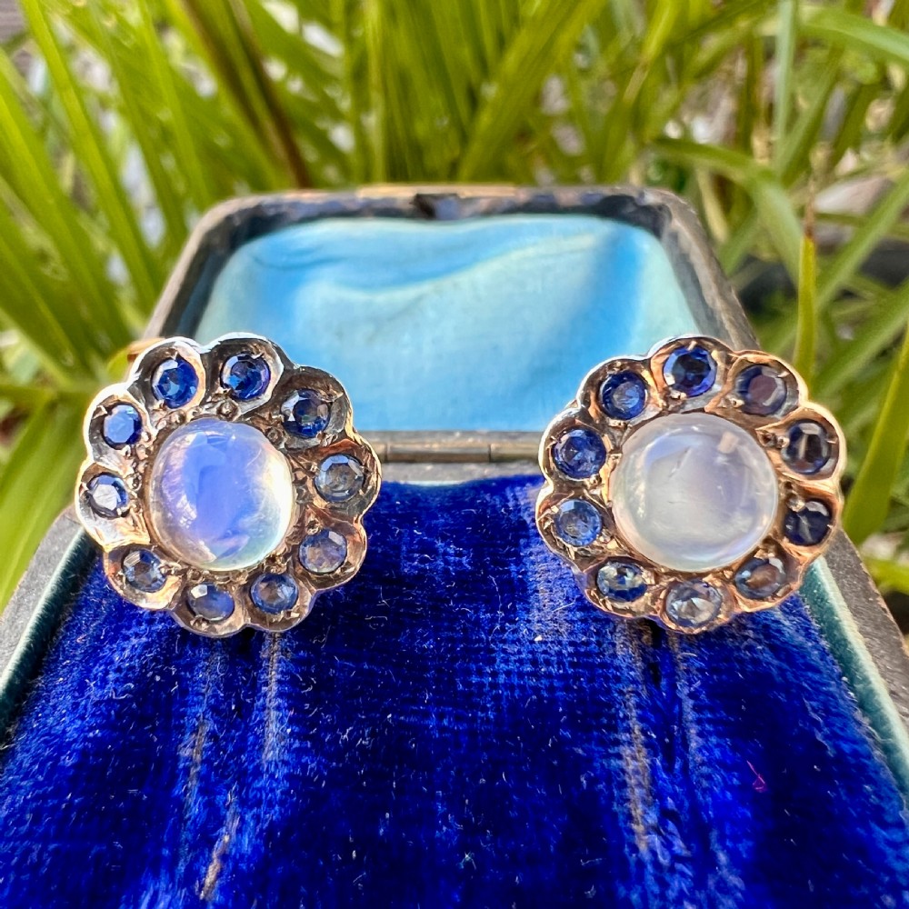 A pair of circular gold screwback earrings, each featuring a round cabochon moonstone surrounded by a halo of small blue sapphires. View X