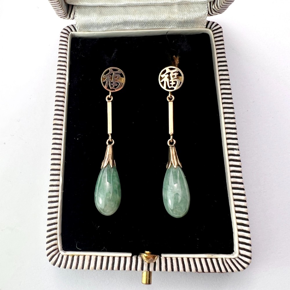 A pair of gold drop earrings featuring circular tops with pierced Chinese characters, slender gold bar links, and teardrop-shaped green jade stones.