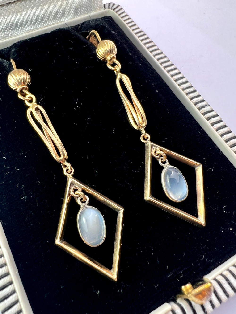 A pair of yellow gold drop earrings featuring ribbed shepherd hooks, linear twisted wire links, and open diamond-shaped frames containing oval moonstone cabochons. View V