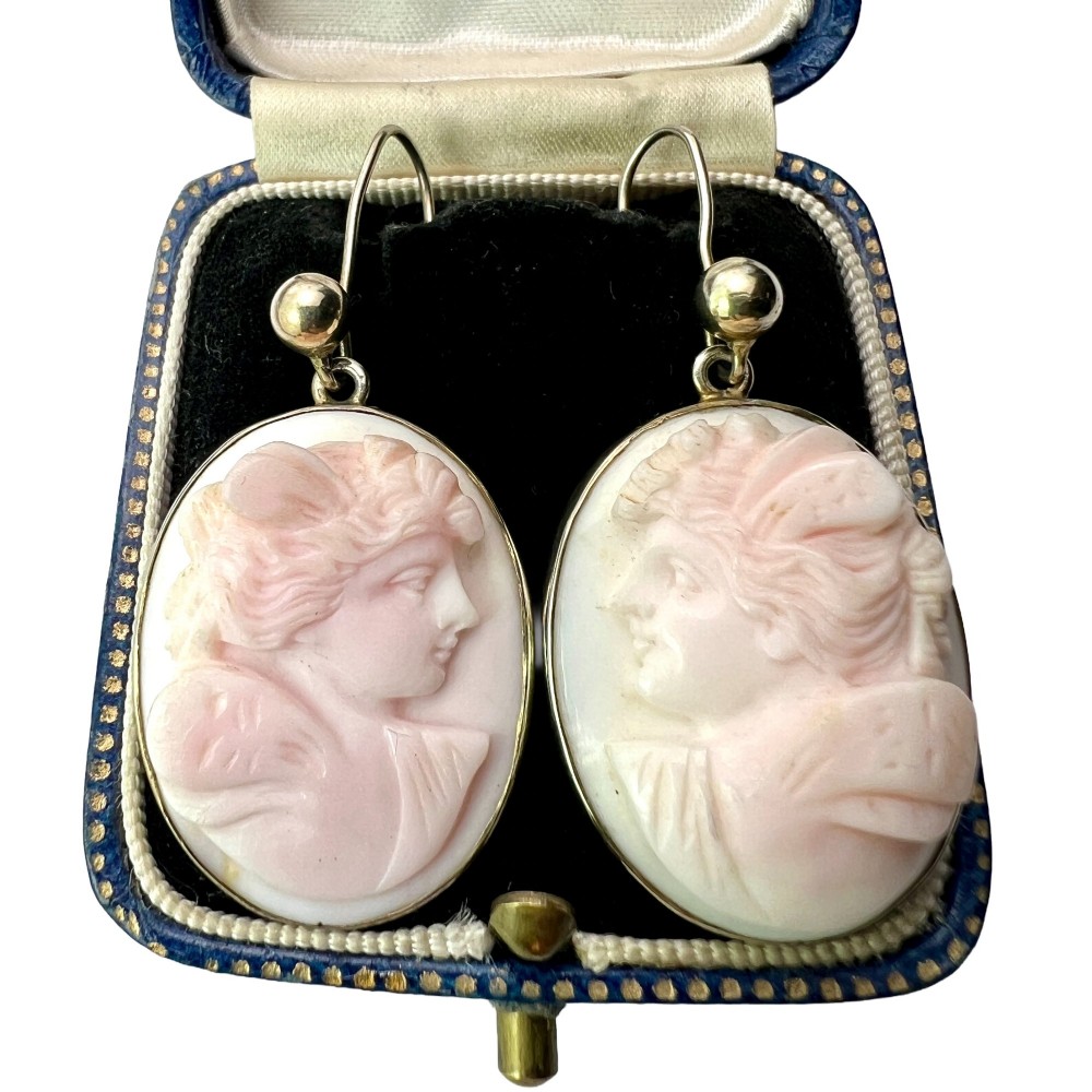 A pair of oval drop earrings featuring carved pink and white queen conch shell cameos depicting classical profile portraits set in silver mounts.