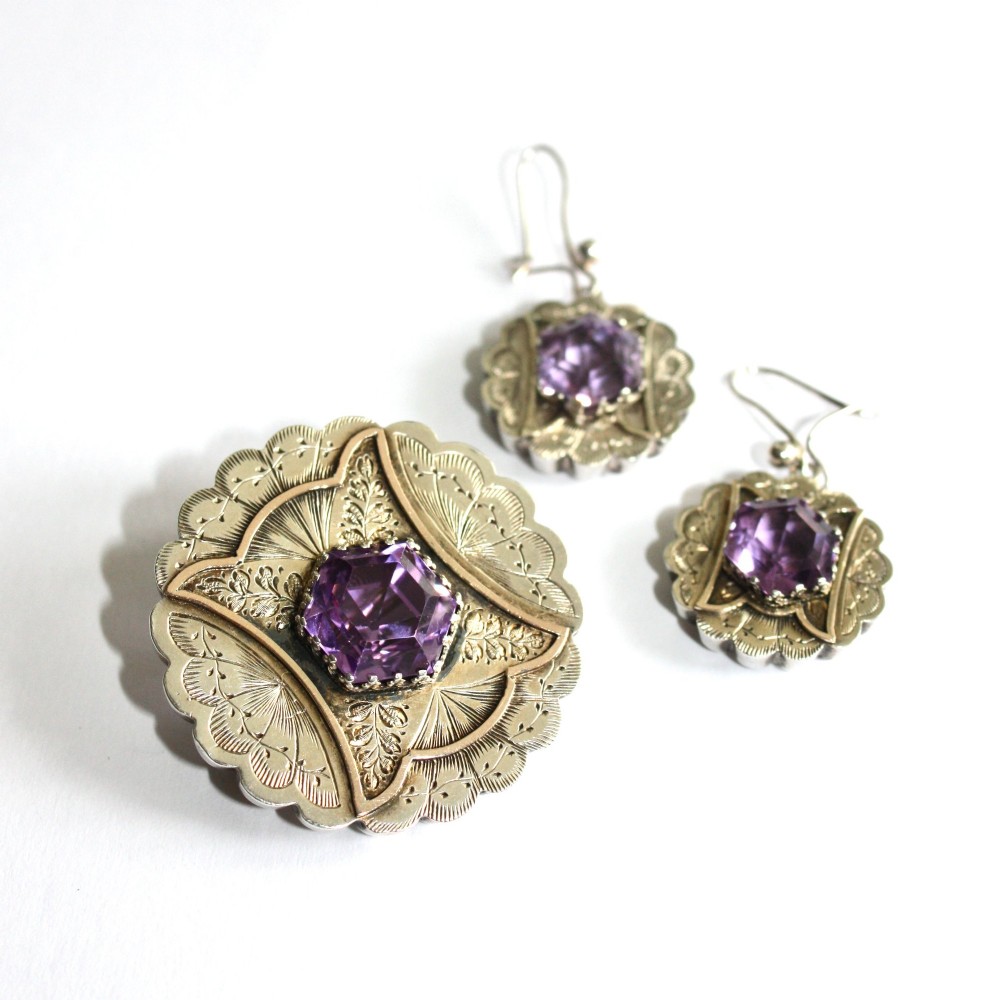 A Victorian silver jewellery set comprising a circular brooch and matching drop earrings, each featuring engraved detailing and a central faceted purple amethyst stone.