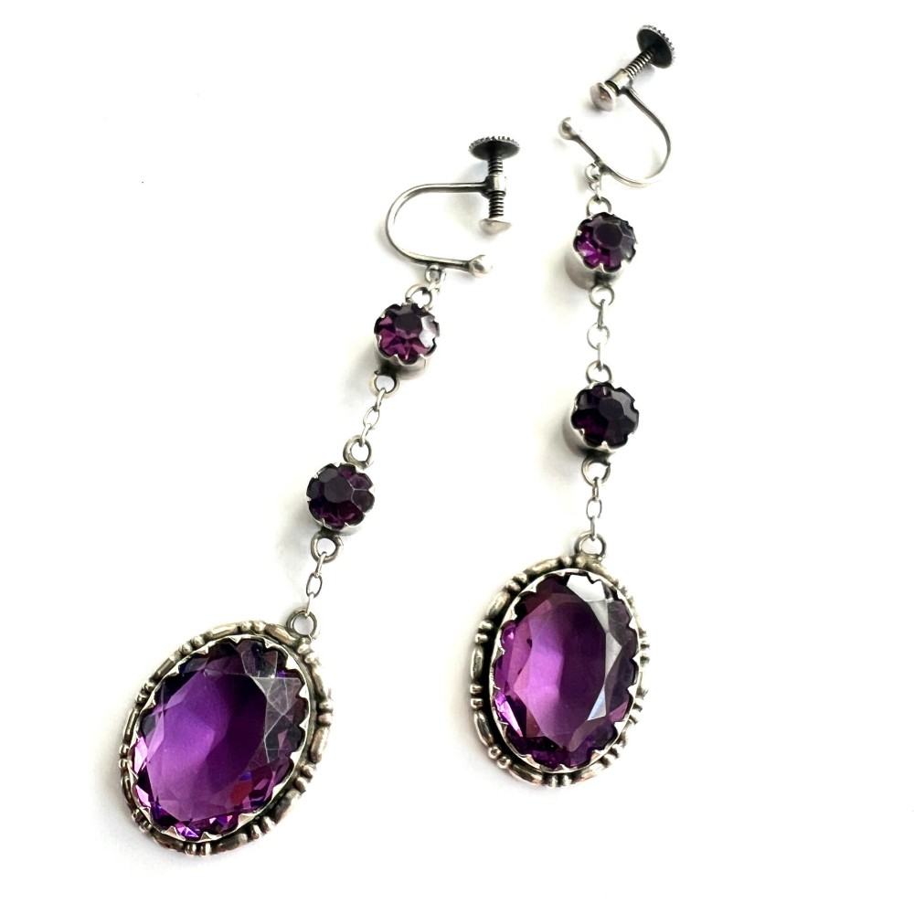 A pair of Art Deco drop earrings featuring faceted purple paste stones set in silver with screwback mechanisms.