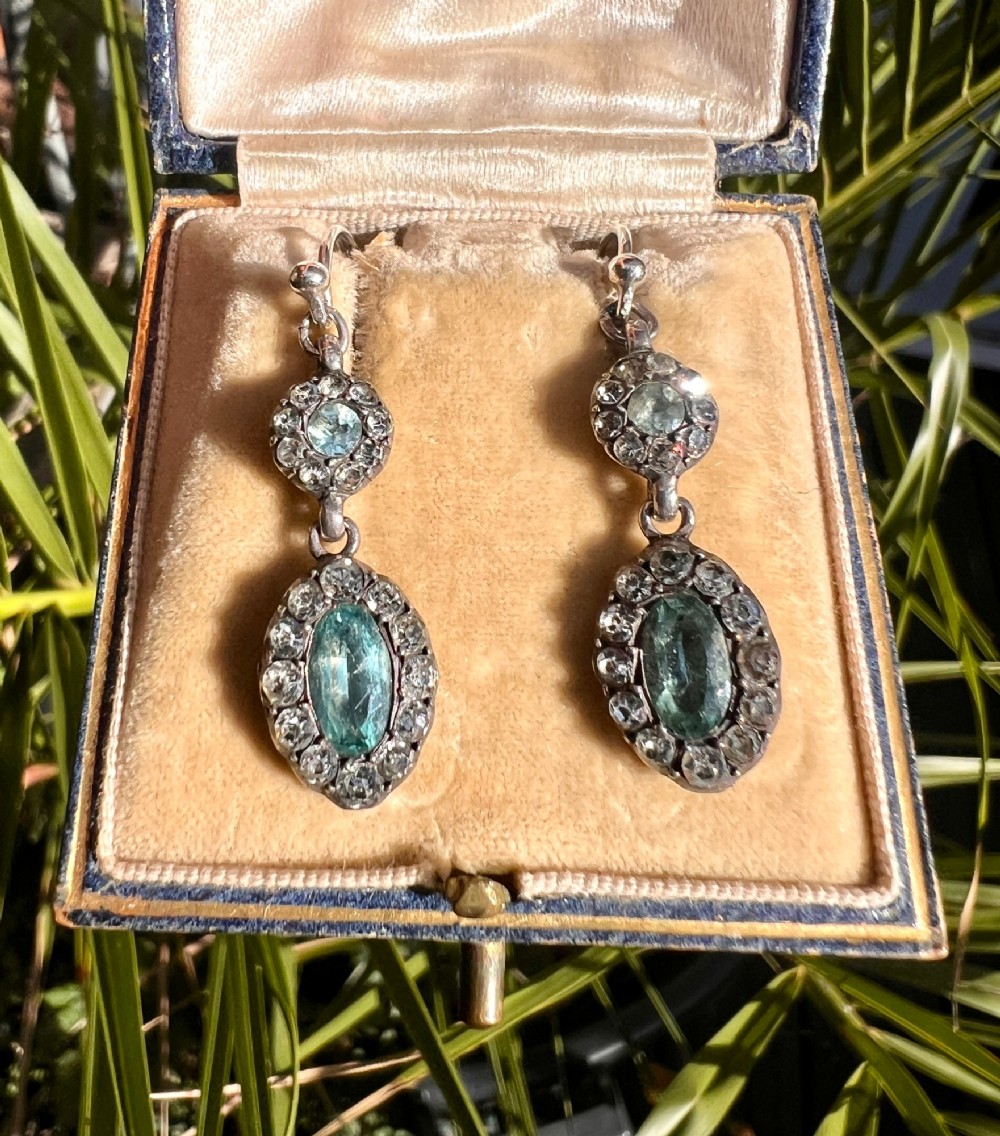 A pair of antique silver drop earrings featuring oval light blue paste central stones surrounded by clear paste halos suspended from wire hooks. View IV