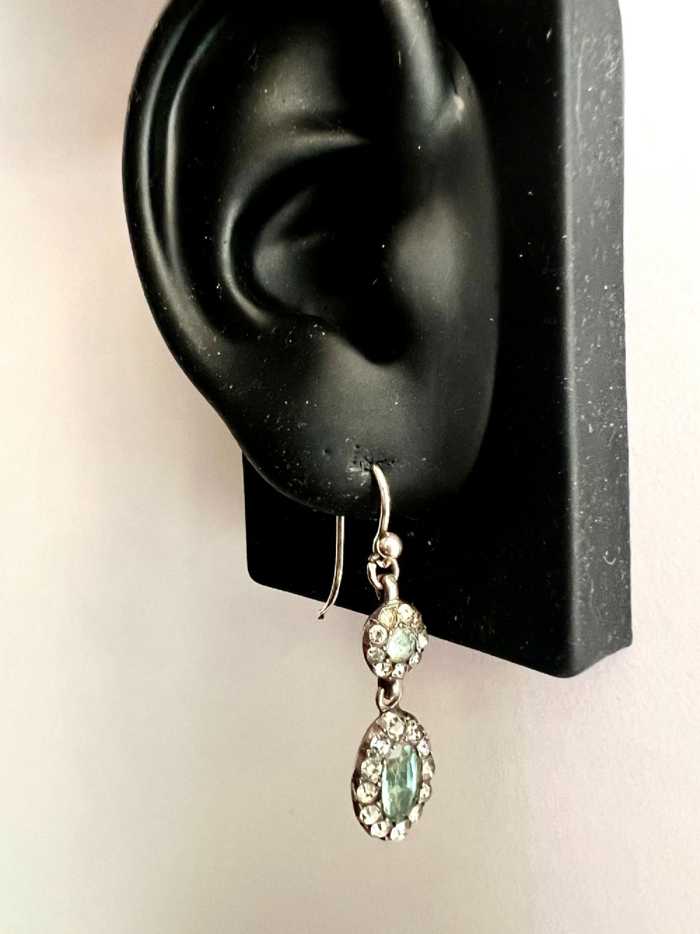 A pair of antique silver drop earrings featuring oval light blue paste central stones surrounded by clear paste halos suspended from wire hooks. View V