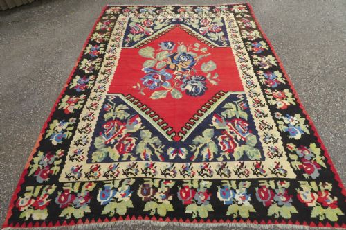 fig 1. An antique flatweave wool rug featuring a central red panel with blue floral motifs, bordered by dark navy and cream botanical patterns.