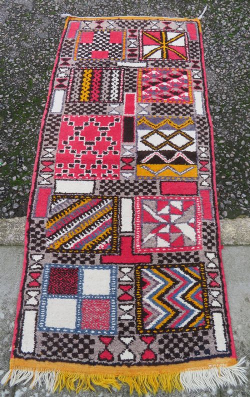 fig 1. A rectangular hand-knotted Moroccan Berber wool rug featuring geometric patchwork panels in pink, yellow, black, grey, and white with fringed ends.