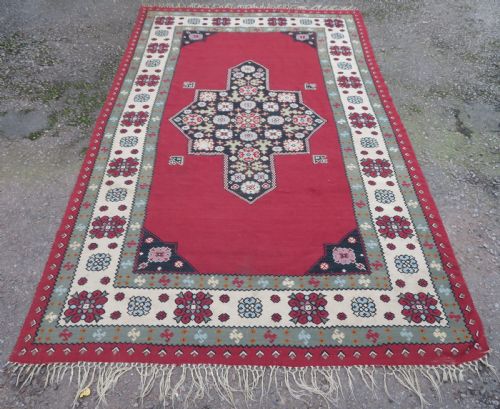 fig 1. An antique flatweave Bessarabian wool rug featuring a red field with a central black and cream floral medallion, surrounded by a wide floral border and fringed edges.