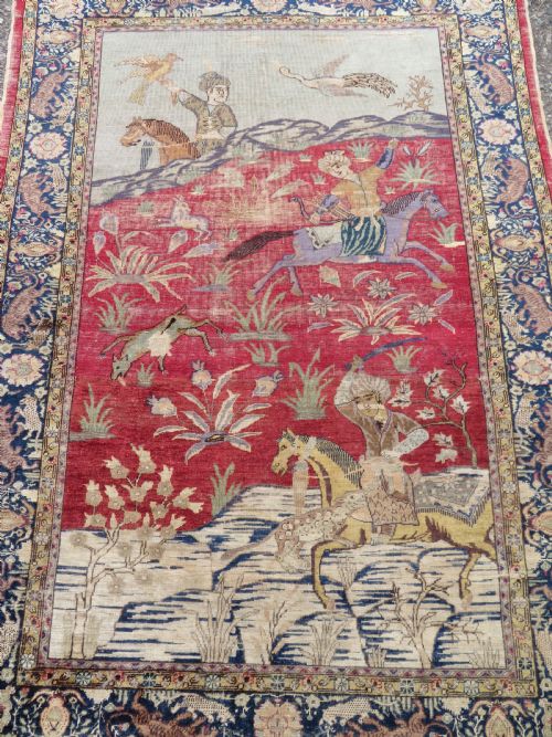 fig 1. A 19th-century West Anatolian silk rug featuring a red central panel depicting horseback hunters, surrounded by a floral and animal motif navy border.