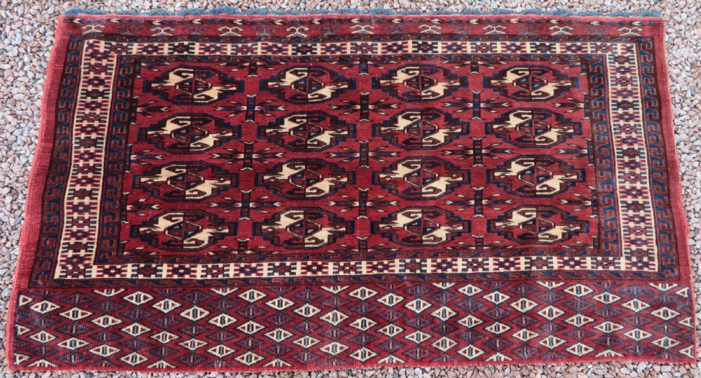 A rectangular hand-knotted Yomut Turkmen chuval rug featuring repeating gul motifs on a deep red field with a geometric border and diamond-patterned lower elem.