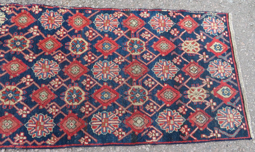 A long, narrow hand-knotted wool runner featuring a repeating geometric Mina Khana floral lattice pattern in navy blue, terracotta red, and cream tones. View V